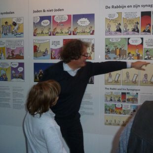 Foto des Karikaturisten Ben Gershom, von der Seite. Im Hintergrund ist die Ausstellung zu sehen, Gerschom erklärt und zeigt auf eines der Bilder.