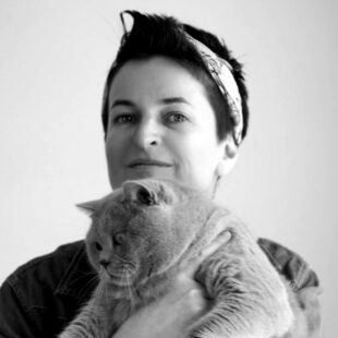 Porträt-Foto der Illustratorin Ira Ginzburg in schwarzweiß. Ginzburg trägt ein Haarband in den kurzen dunklen Haaren und hat eine Katze auf dem Arm