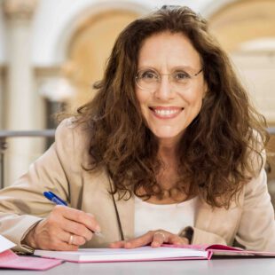 Porträt-Foto der Verlegerin Myriam Halberstam am Tisch sitzend mit braunen langen Haaren und randloser Brille, die von ihr herausgegebene Antologie "#Antisemitismus für Anfänger" signierend.